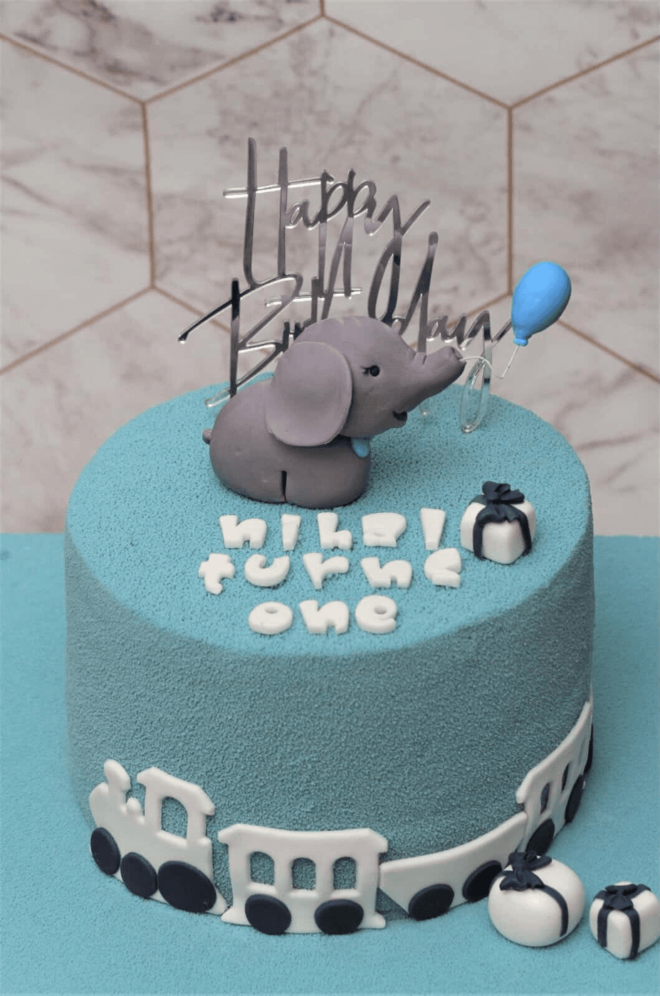 Pale blue velvet cake on a matching blue board, with a modelled grey elephant holding a blue balloon and a mirrored silver "Happy Birthday" topper on top, white fondant letters across the top spelling out a child’s name and "turns one", small white parcels tied with navy ribbon, and a white fondant train with navy wheels running right around the base