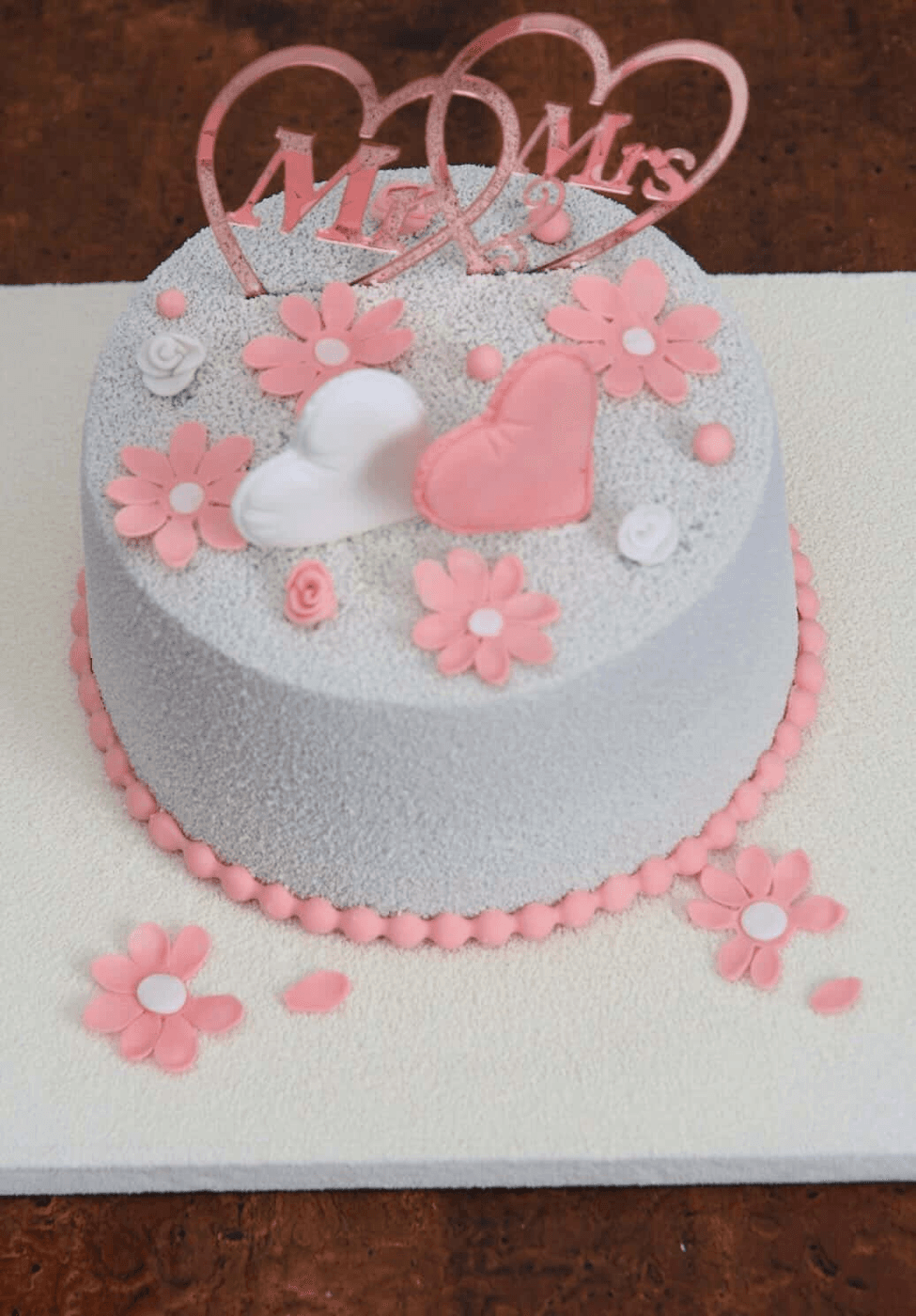 Pale grey velvet cake dressed with pink fondant daisies, one white and one pink sculpted heart, two small white fondant roses and one small pink one, finished with a piped pink pearl border around the base and a rose-gold mirrored Mr & Mrs topper of two interlocking hearts, with loose daisies and petals scattered on the white board