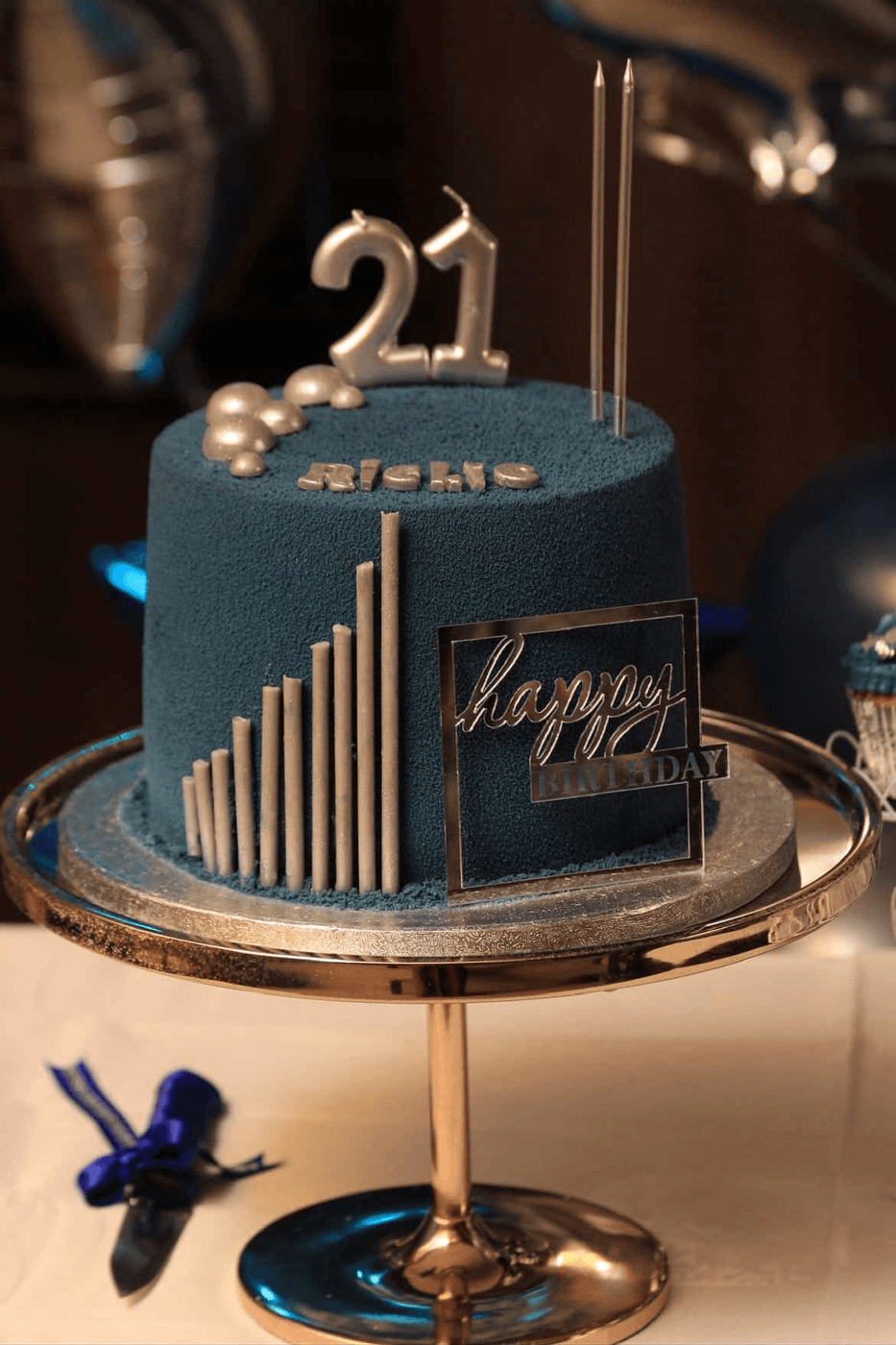 Deep teal-blue velvet cake on a mirrored rose-gold pedestal stand, with a silver number 21 candle and a cluster of silver pearls on top, two tall thin silver candles behind, the birthday guest’s name spelled out in small silver letters across the front, a graduated fan of silver bars rising up one side and a mirrored acrylic plaque reading "happy birthday"