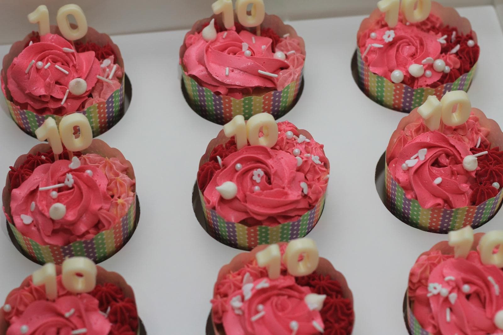 Nine rose-swirl cupcakes in striped paper wraps, piped in pink and deep red buttercream and scattered with white sugar pearls and blossoms, each carrying a white chocolate number 10